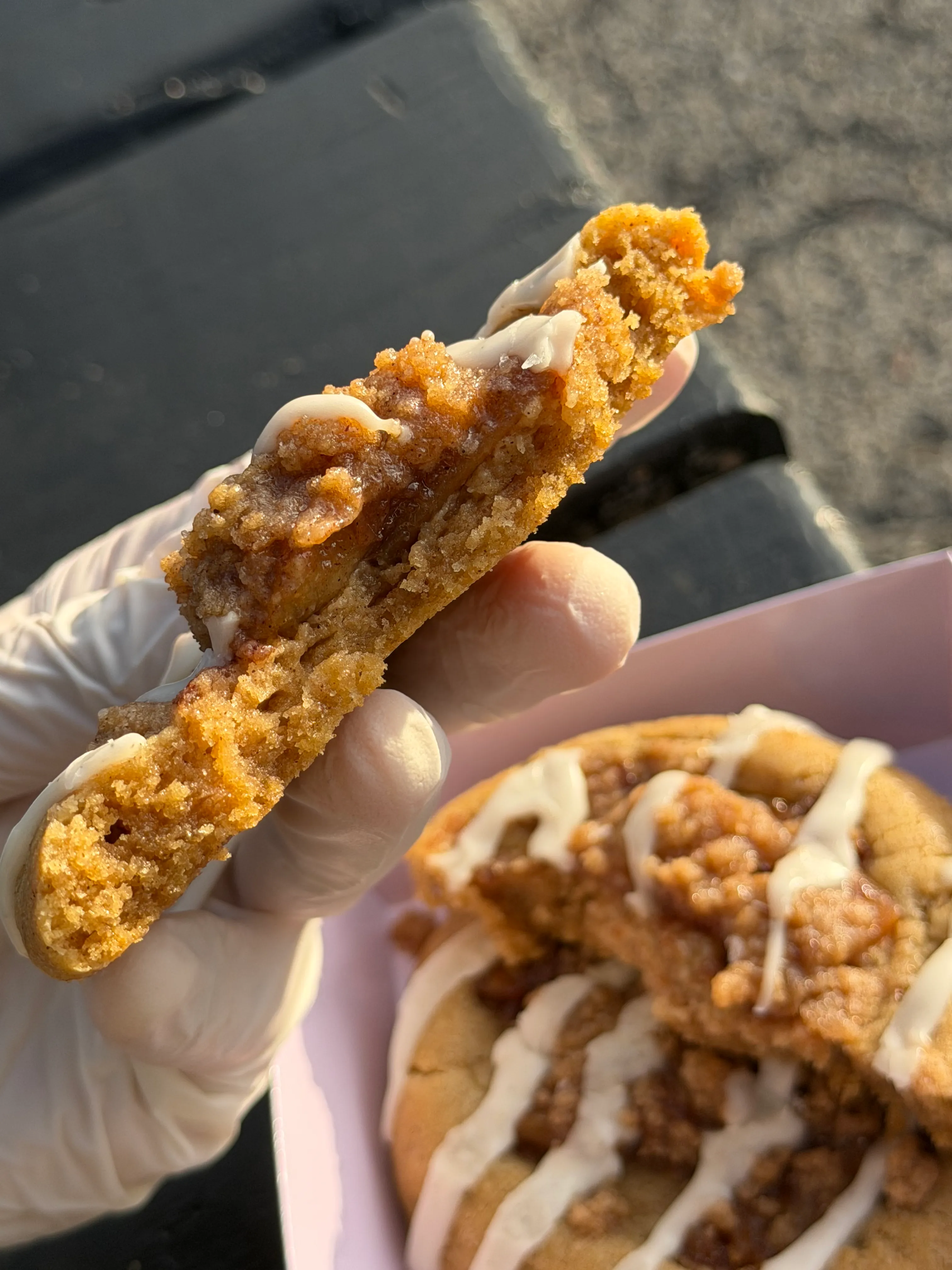 Gloved hand holding a thick crumb-topped cookie above a lavender bakery box.