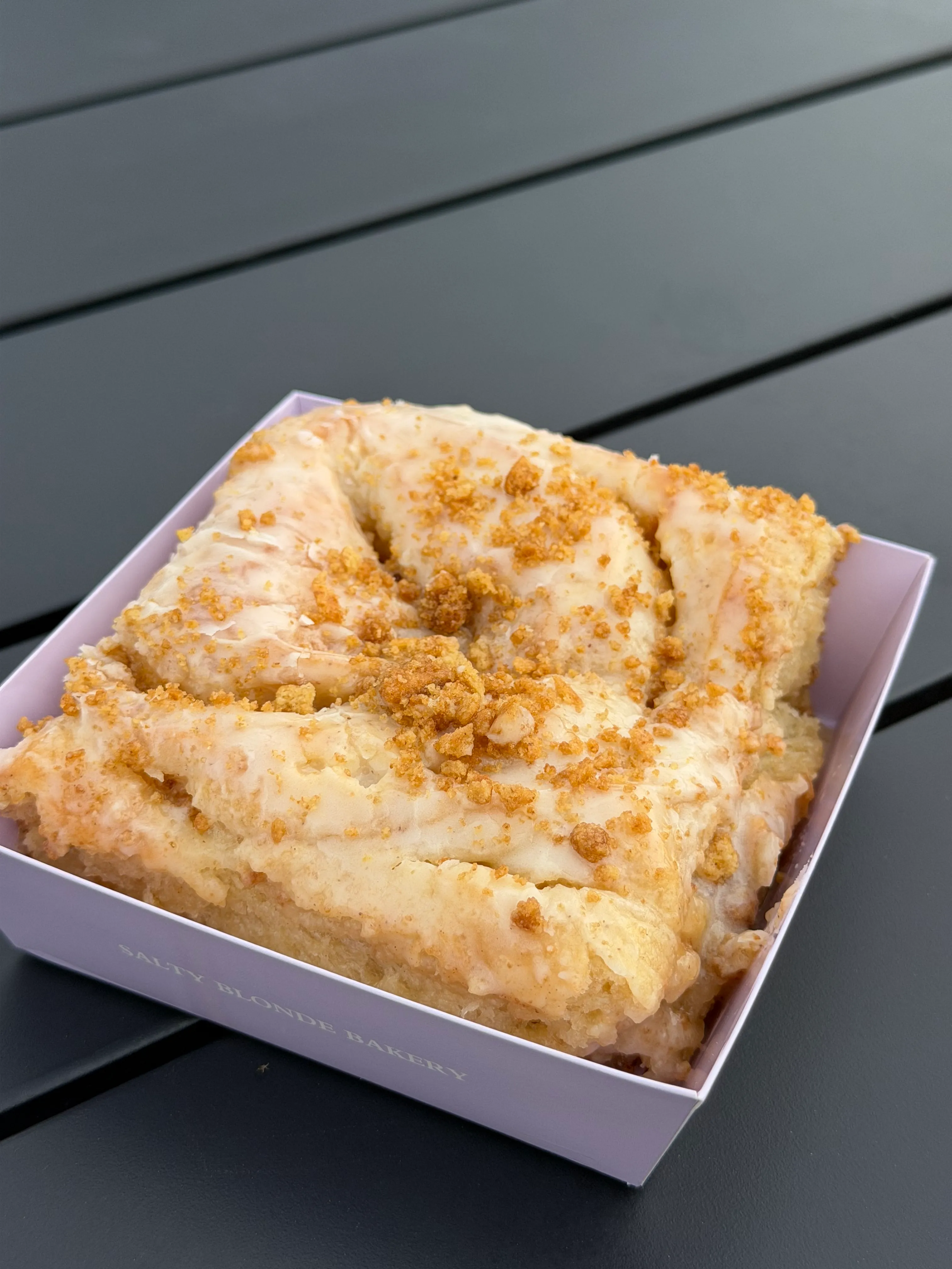 Large glazed bakery roll in a lavender Salty Blonde Bakery box on a dark outdoor table.