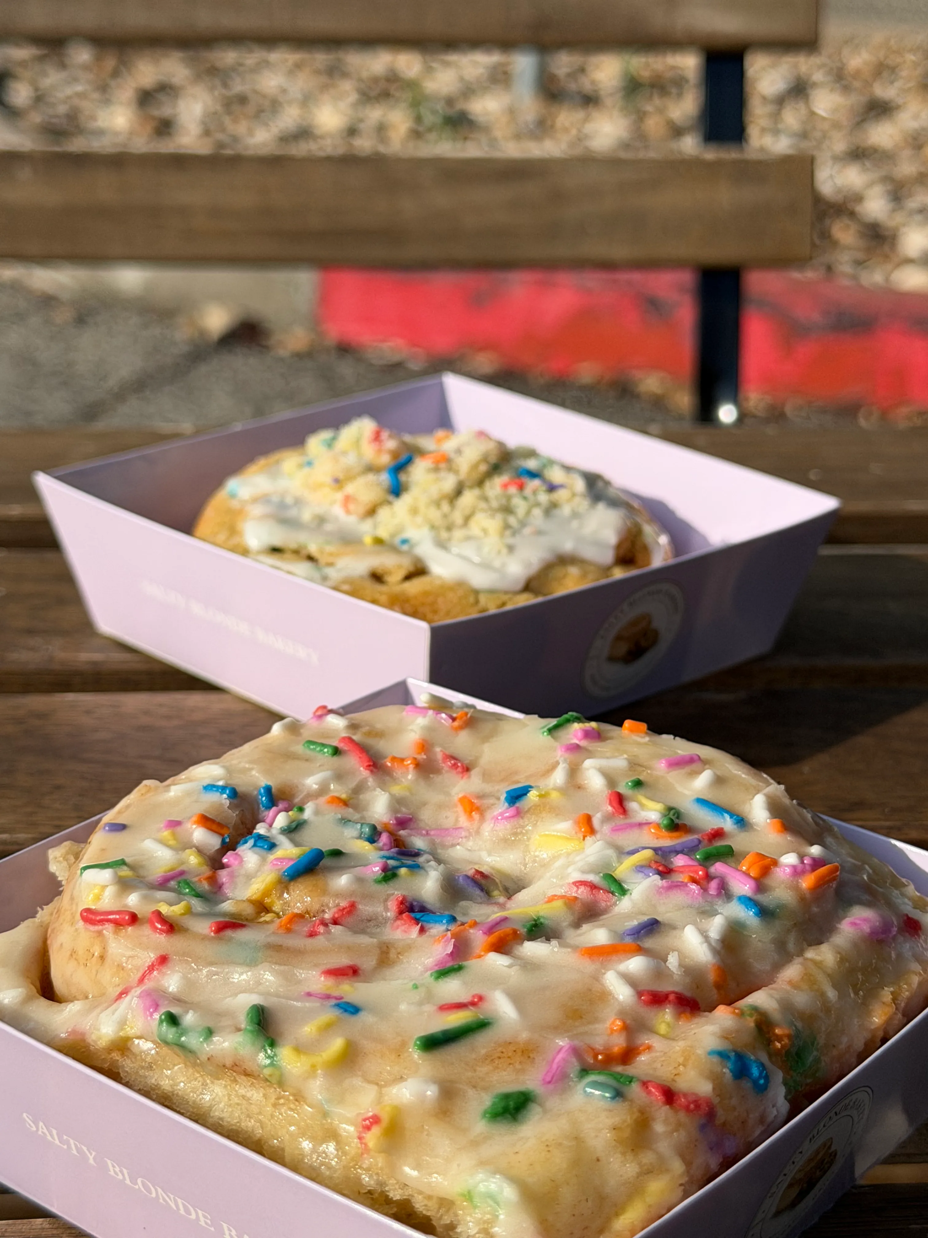 Colorful frosted bakery specials with rainbow sprinkles arranged in lavender boxes outdoors.