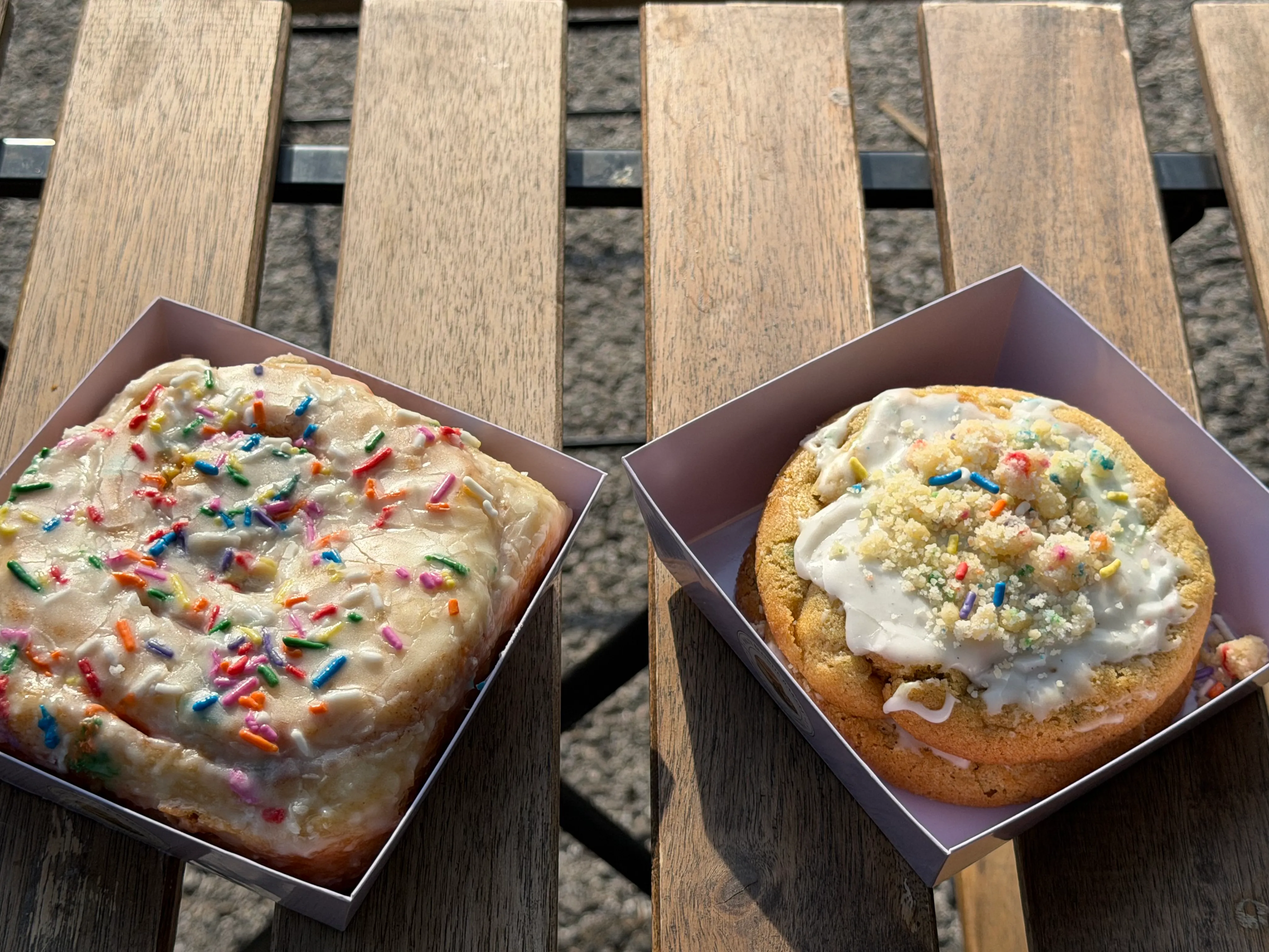 Two colorful frosted bakery specials in lavender boxes on an outdoor wooden table.