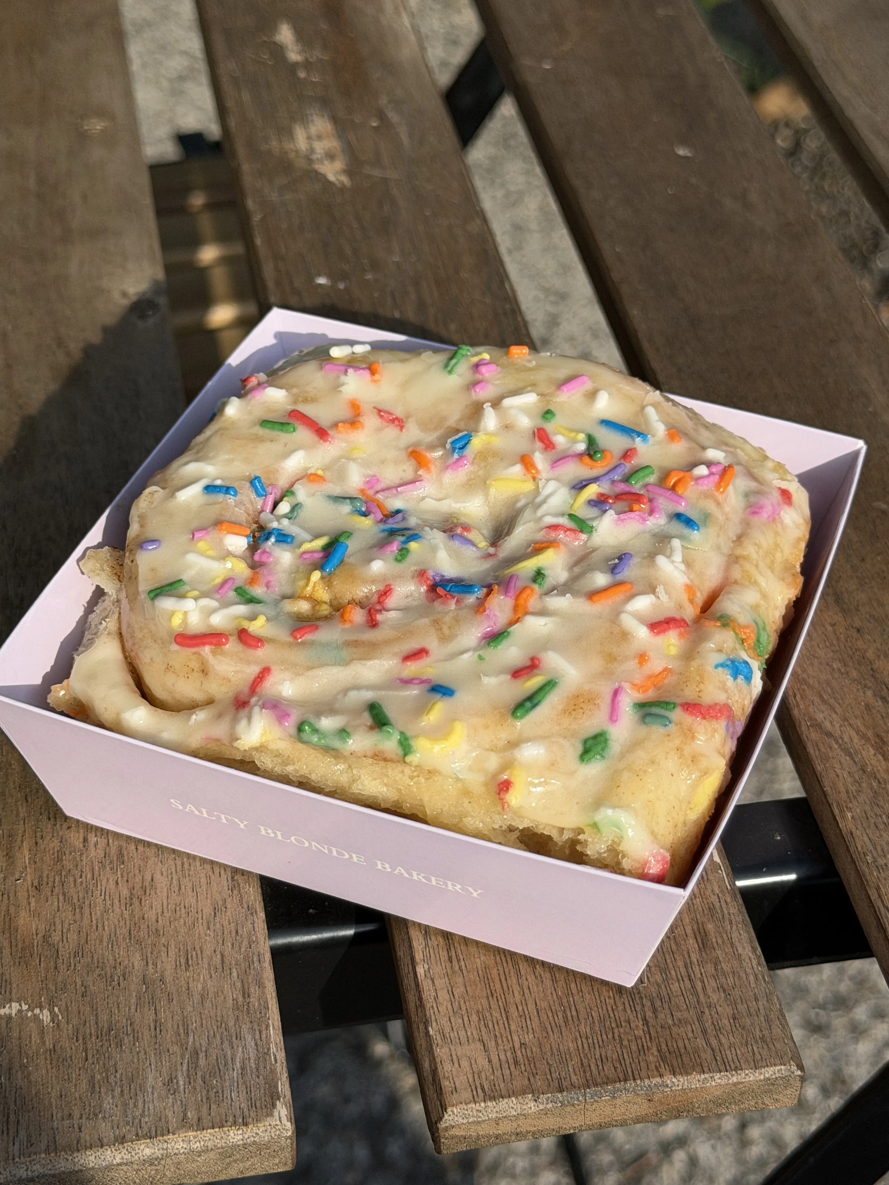 Large frosted sprinkle roll in a lavender bakery box on a wooden picnic table.